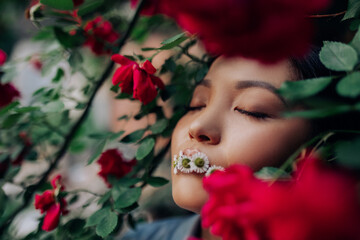 Naklejka premium Portrait of young woman with chamomile flowers in her mouth among red rose.