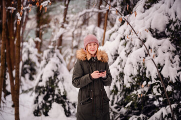 Young blonde girl in winter outfit stands in snowy park using smartphone