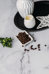 Coffee beans in a white saucer on a marble background next to a black tray greens milk jug decorative star