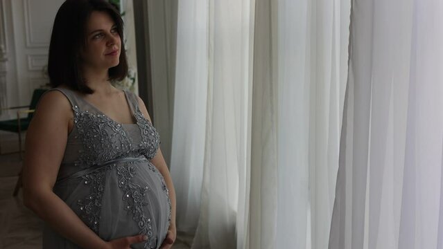 Young pretty pregnant girl in beautiful smart dress standing in porsch apartment. Pregnant woman with short hair smiling and touching her belly. Mother-to-be, future mother, motherhood