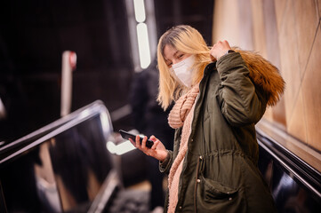 Blonde young woman in winter outfit and protective mask using smartphone going up the escalator