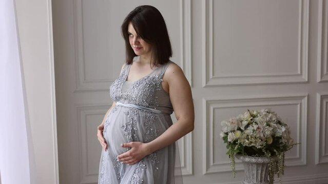 Young pretty pregnant girl in beautiful smart dress standing in porsch apartment. Pregnant woman with short hair smiling and touching her belly. Mother-to-be, future mother, motherhood