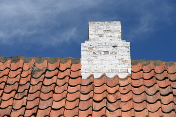 Rustikales Hausdach mit roten handgestrichenen Dachpfannen und weißem Schornstein vor blauem Himmel