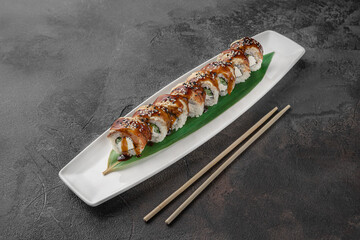 set of philadelphia roll with eel and green bamboo leaf in a white ceramic plate with chopstick on a dark gray textured background, side view