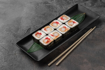 set of square maki rolls with tiger shrim, caviar and green bamboo leaf in a black ceramic plate with chopstick on a dark gray textured background, side view