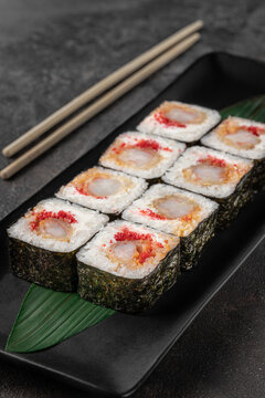 set of square maki rolls with tiger shrim, caviar and green bamboo leaf in a black ceramic plate with chopstick on a dark gray textured background, side view, close-up