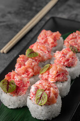 set of rolls with salmon, red flying fish caviar and green bamboo leaf in a black ceramic plate with chopstick on a dark gray textured background, side view, close-up