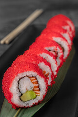 set of philadelphia rolls with red flying fish caviar, avocado, salmon and green bamboo leaf in a black ceramic plate with chopstick on a dark gray textured background, side view, close-up