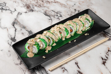 set of maki rolls with chuka wakame, salmon, nori, sesame sauce with nuts and green bamboo leaf in a black ceramic plate with chopstick on a bright white textured marble background, side view