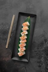 set of maki rolls with tiger shrimp, salmon, microgreens and green bamboo leaf in a black ceramic plate with chopstick on a dark gray textured background, top view
