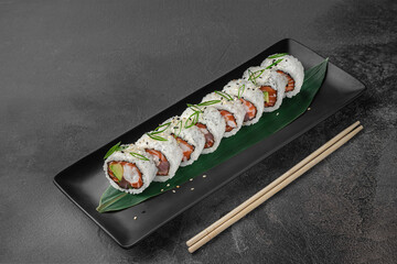 set of maki rolls with salmon tuna, shrimp, avocado, rice and green bamboo leaf in a black ceramic plate with chopstick on a dark gray textured background, side view