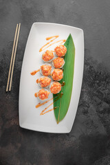set of hot baked rolls with salmon, rice, avocado, chili sauce and green bamboo leaf in a white ceramic plate with chopstick on a dark gray textured background, top view