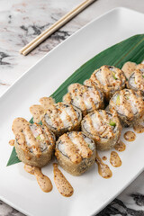 set of fried rolls with tiger shrimp, avocado, sesame sauce and green bamboo leaf in a white ceramic plate with chopstick on a bright textured marble background, side view, close-up
