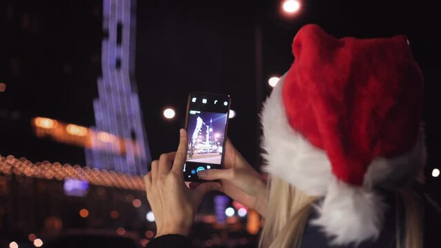 A smiling woman in a Santa Claus hat taking a photo of the night city with her smartphone on Christmas eve. A happy woman takes pictures on her mobile phone on the eve of the Christmas holidays.