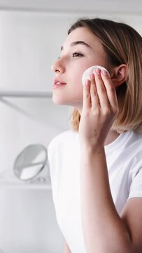Facial hygiene. Skin care. Freshness beauty. Morning routine. Relaxed woman with nude makeup cleansing flawless face using cotton pad tonic on light vertical background.