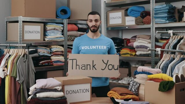 Portrait view of the caucasian volunteer man holding cupboard banner with thank you phrase and looking at the camera with smile. Shelves with belongings at the background. Humanitarian aid concept