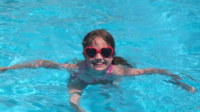 Smiling cute little girl in heart shaped sunglasses in swimming pool on sunny day. Childhood summer vacation concept. Happy kid playing. Healthy outdoor sport activity for children. Kids beach fun