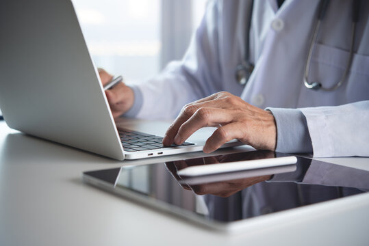 Male Doctor Working On Laptop Computer In Medical Office With Digital Tablet And Medical Chart On Table