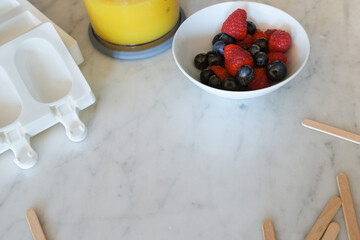 Preparation of healthy sugar-, lactose-, gluten-free summer treat: homemade mango popsicles (ice lolly) with blueberry and raspberry. Silicon forms, wooden sticks on marble background. Copy space.