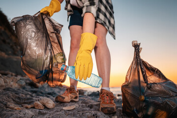 Female volunteer holding two polyethylene bags and picks up a plastic bottle on the beach. Close up of hand. Low angle view. The concept of cleaning the coastal zone