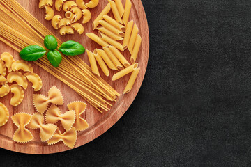 Spaghetti, farfalle, rigatoni, maccheroni, penne rigate, cresta di gallo, raw, basil, on wooden brown plank tabletop background, top view, space to copy text.