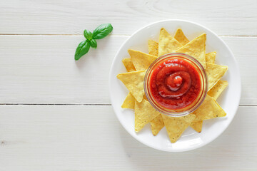 Nachos, with tomato salsa, leaf of green basil, on plate, on white wooden board background, top view, space to copy text..