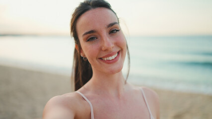 Young athletic woman with long ponytail wearing beige sports top smiling makes selfie on the sea...