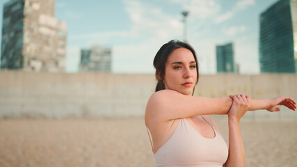 oung athletic woman with long ponytail wearing beige sports top is stretching on the beach on modern city background.