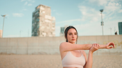 oung athletic woman with long ponytail wearing beige sports top is stretching on the beach on modern city background.