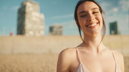 Young athletic woman with long ponytail wearing beige sports top walks along the beach at dawn on modern city background