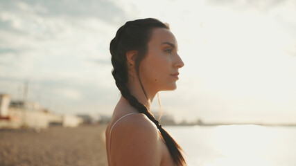 Young athletic woman with braided pigtails wearing beige sports top walks along the beach at dawn...