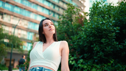 Young woman with freckles and dark loose hair wearing white top and jeans is enjoying the view of modern city. Girl with long eyelashes sits in public garden on modern building background