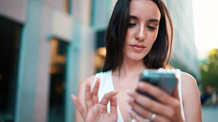 Beautiful woman with freckles and dark loose hair wearing white top is walking down the street with smartphone in her hands. Girl uses mobile phone on modern city background