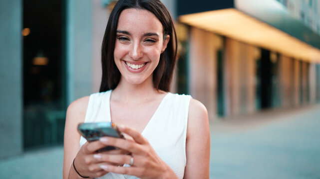 Beautiful Woman With Freckles And Dark Loose Hair Wearing White Top Is Walking Down The Street With Smartphone In Her Hands. Girl Uses Mobile Phone On Modern City Background