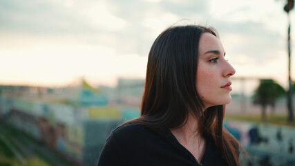 Obraz premium Portrait of cute girl with freckles and loose brown hair wearing black sports hoodie looks at the camera and walks on the bridge of the modern city background