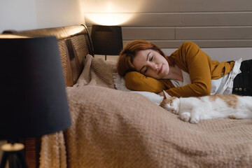 Middle age woman holding cute ginger cat. Female hugging her lovely short hair kitty in bedroom in the evening. Copy space. Adorable domestic pet concept.