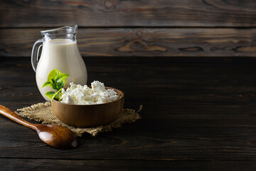 Homemade cottage cheese in a wooden bowl on a canvas napkin. A jug of milk in the background. A wooden spoon on a hundred. Wooden background, space for text