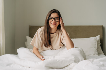 Portrait of a young twenties female student brunette in glasses sitting on the bed.