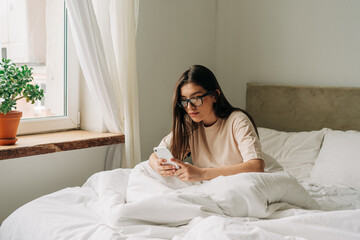 A young twenties caucasian woman sitting on a bed by the window in a bright bedroom uses a phone