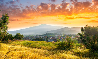Obraz premium view from hill with golden grass and green bushes to a valley town with majectic mountains and scenic cloudy sunset on background