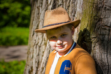 Cute boy posing in a cowboy hat in the woods by a tree. The sun's rays envelop the space....