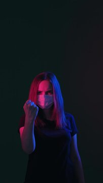 Threaten gesture. Covid-19 restriction. Angry woman in protective mask neon light glow showing fist looking at camera isolated on black vertical. Quarantine measures. Aggressive emotion. New normal
