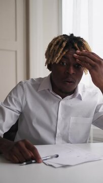 Shocked news. Appalled black man. Work problem. Surprised guy sitting desk expressing bewildered emotion in light office room interior vertical.