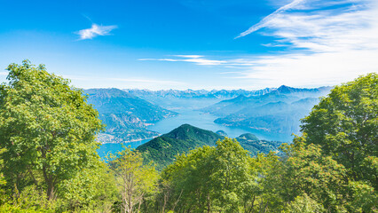 Fototapeta premium Aerial view of Lake Como, Lombardy, Italy, from a viewpoint on the italian alps. Green trees in the foreground. Blue sky with white clouds on the background.