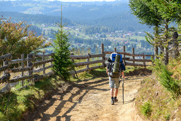A traveler with a large backpack walks along a country road with a wooden fence in a mountainous area overlooking a valley with small houses, rear view