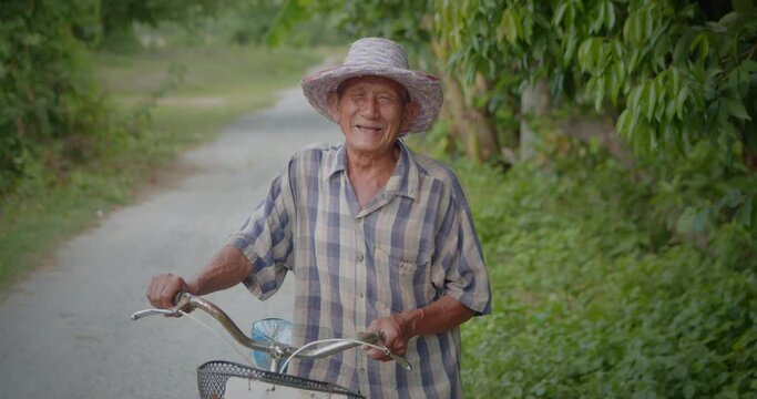 Slow motion scene of happy smiling and laughing Asian man who is poor elderly villager, older than seventy stands wearing old hat leading the bicycle on the roadside of his village.