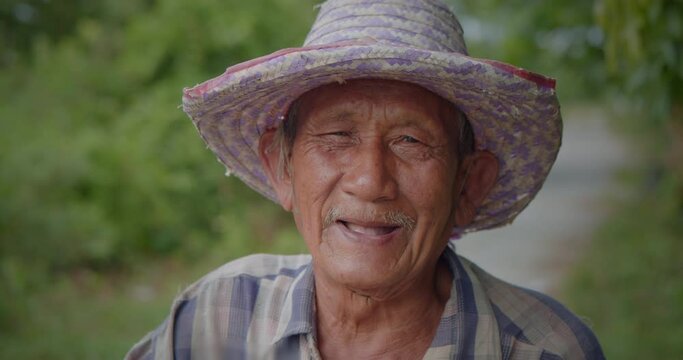 Slow motion scene of happy smiling and laughing Asian man who is poor elderly villager, older than seventy stands wearing old hat leading the bicycle on the roadside of his village.