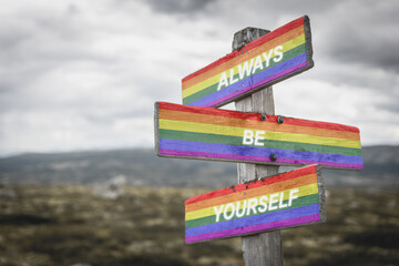 Pride flag on wooden signpost outdoors in nature with the text quote always be yourself. Lgbtq and equality concept.