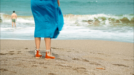 Legs of a European girl in a blue dress walking in shoes on the sand on a beach