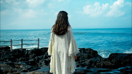 Fototapeta na wymiar A young European girl with long black hair and a white robe walks to Water blow with cliffs and strong waves of seawater with clouds and blue sea